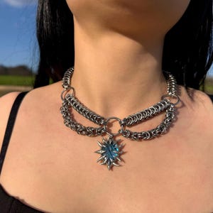 May include: A silver chainmail necklace with a sunburst pendant featuring a blue gemstone. The necklace has multiple layers of chain and a large central ring. The pendant is a starburst shape with pointed edges.