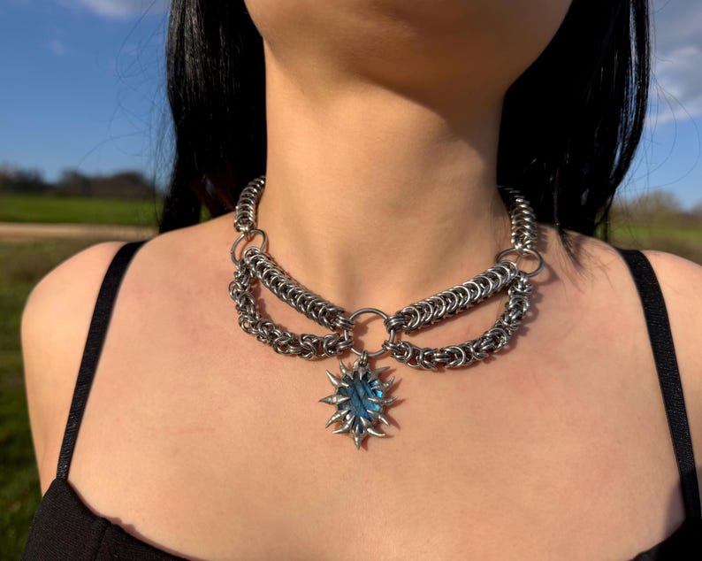 May include: A silver chainmail necklace with a sunburst pendant featuring a blue gemstone. The necklace has multiple layers of chain and a large central ring. The pendant is a starburst shape with pointed edges.