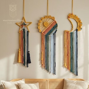Op de afbeelding: Drie macramé wandhangers in de vorm van een ster, een zon en een maansikkel. Elk stuk is gemaakt met gele koordframes en veelkleurige hangende draden in tinten geel, oranje, blauwgroen en marineblauw. De tekst macrameablom is zichtbaar.
