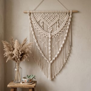 May include: A cream macrame wall hanging with detailed woven patterns and a wooden dowel. The design includes geometric shapes and cascading fringe, creating a bohemian style. The wall decor is displayed against a neutral-toned wall, adding a touch of texture.