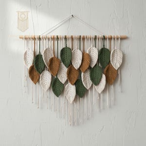 Puede incluir: Un tapiz de pared de macramé con una barra de madera y una serie de hojas hechas a mano en tonos blanco, verde y marrón. Las hojas están suspendidas por cuerdas, creando un diseño en capas y texturizado. Una pieza de macramé más pequeña cuelga arriba.