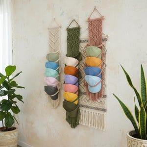 May include: Three macrame hat organizers in cream, olive green, and dusty rose hang on a wall. Each holds several baseball caps in various colors, including blue, pink, and yellow. The organizers have a bohemian style with fringe and wooden dowels.