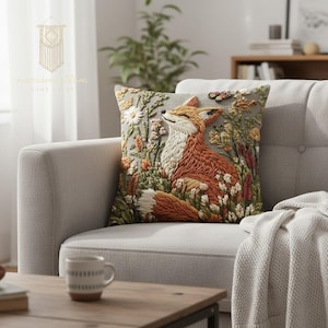 Puede incluir: Cojín cuadrado bordado con un zorro en un campo de flores. El zorro tiene pelaje naranja y blanco, con una expresión serena. El cojín está sobre un sofá gris, con una manta blanca y una mesa de madera con una taza.