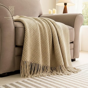 May include: A beige textured throw blanket with fringe detail draped over a gray armchair. The blanket has a woven pattern and is made of a soft material. The scene includes a lamp and a patterned rug.