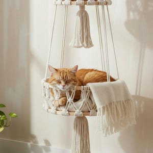 May include: An orange tabby cat sleeps peacefully in a macrame cat bed. The bed is suspended from a wooden frame with woven rope and a cream tassel. A cream blanket is draped over the edge of the bed, creating a cozy space.