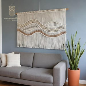 Peut inclure: Une tenture murale en macramé crème et beige avec un motif de vagues. La décoration murale est suspendue à une cheville en bois et présente des nœuds et des franges complexes. Le texte "macrameablom HOME DECOR" est visible. La tenture mesure environ 100 cm de large.