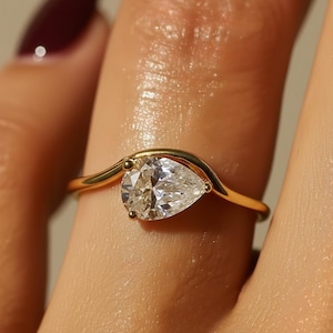 May include: A gold ring featuring a pear-shaped diamond. The ring's band curves around the finger, with the diamond set in a prong setting. The ring is shown on a finger with a dark red nail polish.