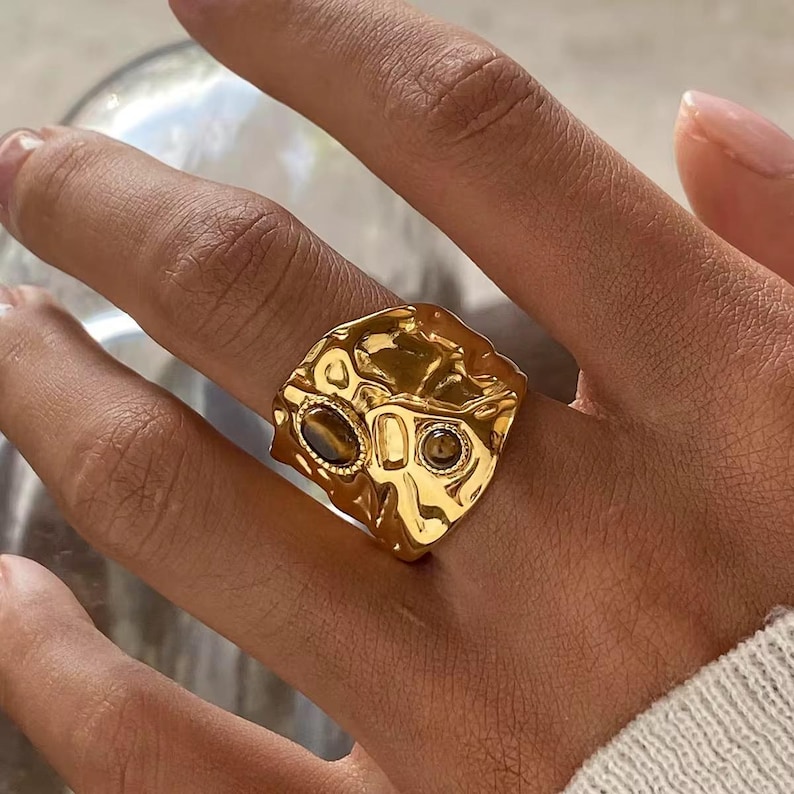 May include: A gold-colored ring with a textured surface and two oval-shaped, dark brown gemstones. The ring is worn on a finger, showcasing its unique, abstract design. The ring's design is reminiscent of a hammered metal style.18k Gold Plated Ring for Women, Gold PVD Stainless Steel Ring, Non-Tarnishing Ring, Waterproof Jewelry For Women, Cute Gift Idea for her