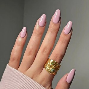 May include: A hand displaying long, oval-shaped nails in a pale pink shade. A gold ring with an intricate pattern and green stone details is on the ring finger. The nails and ring are the main focus against a plain backdrop.