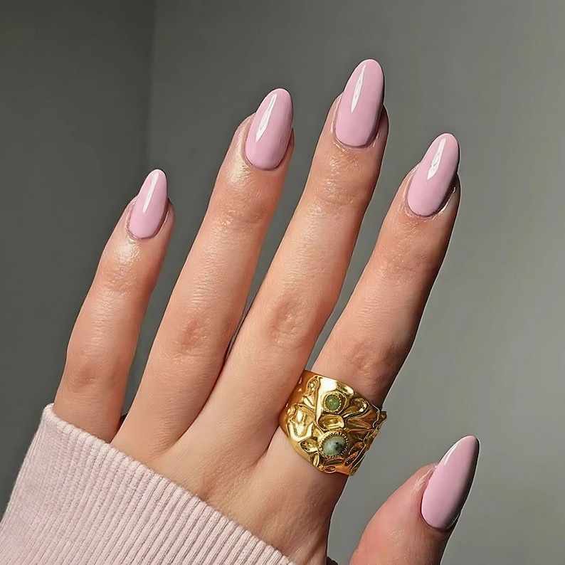 May include: A hand displaying long, oval-shaped nails in a pale pink shade. A gold ring with an intricate pattern and green stone details is on the ring finger. The nails and ring are the main focus against a plain backdrop.