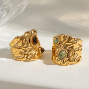 May include: Two gold-coloured statement rings with textured surfaces. One ring features brown and amber-coloured stones, while the other has green and teal stones. The rings are set against a white background.