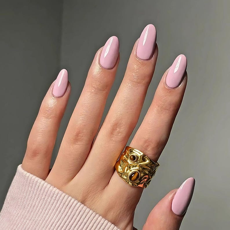 May include: A hand displaying long, oval nails painted a pale pink. A gold ring with a textured design and brown stone accents is on the ring finger. The nails have a glossy finish, set against a neutral backdrop.