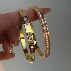 May include: Two gold-colored bangles. One bangle is adorned with rectangular, clear gemstones. The other bangle features a series of rectangular, clear gemstones. The bangles are held up by a person's fingers.