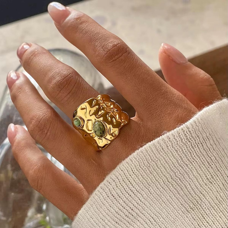 May include: A gold ring with a textured, uneven surface. The ring has two small, round, green gemstones and one oval green gemstone. The ring is worn on a finger.