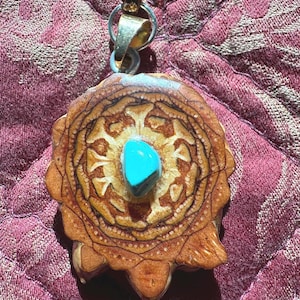 Puede incluir: Colgante artesanal con un diseño detallado de piña y una piedra turquesa central. El colgante está suspendido de un cordón marrón. Los cálidos tonos marrones de la piña contrastan con el azul brillante de la piedra.