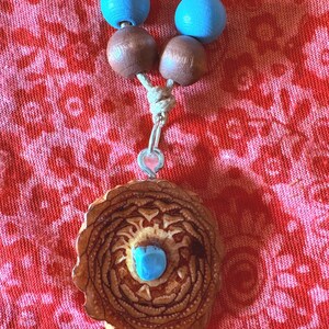 Puede incluir: Un collar hecho a mano con un colgante grande de madera tallada y una piedra turquesa. El collar tiene cuentas de madera marrón y azul, ensartadas en un cordón natural. El fondo es una tela floral roja.