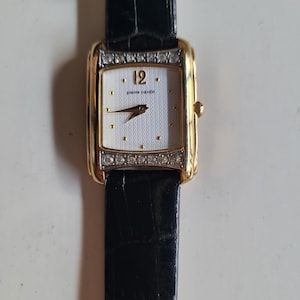Könnte beinhalten: Eine goldfarbene Pierre Cardin Armbanduhr mit rechteckigem Zifferblatt. Die Uhr hat ein weißes Zifferblatt mit goldfarbenen Zeigern und Stundenmarkierungen sowie ein schwarzes Lederarmband. Die Lünette ist mit kleinen, klaren Kristallen besetzt. Das Zifferblatt zeigt die Zahl 12.