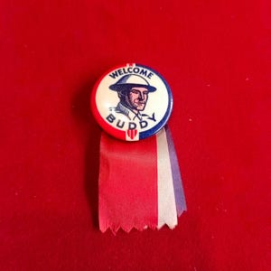 Puede incluir: Un pin vintage con temática patriótica. El pin presenta un soldado con casco, las palabras "WELCOME BUDDY" y cintas rojas, blancas y azules. El pin es circular y tiene un fondo blanco con detalles azules y rojos.