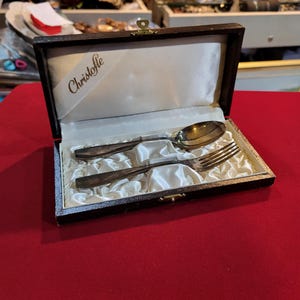 May include: A silver spoon and fork set inside a dark brown box with a cream-colored satin lining. The box has a gold clasp and a white ribbon with the word "Christofle" printed on it. The set is displayed on a red surface.