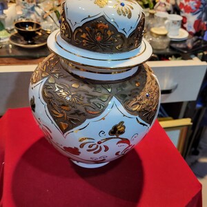 Puede incluir: Un jarrón de cerámica blanco y dorado con tapa. El jarrón presenta diseños florales marrones y dorados ornamentados, acentuados con joyas rojas y azules. La tapa está rematada con un remate dorado, y el jarrón se encuentra sobre una superficie roja.