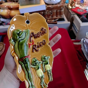 Può includere: Applique da parete in ceramica a forma di foglia con tre rane verdi con pantaloncini gialli e una fascia rossa. Le parole "Puerto Rico" sono scritte in marrone. Lo sfondo è marrone chiaro.