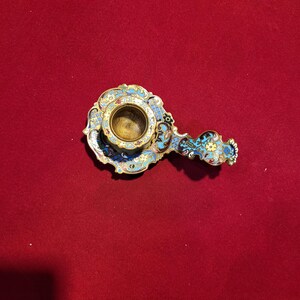 May include: An ornate, antique candle holder with a brass cup and a decorative handle. The candle holder features a colorful enamel design with blue, turquoise, yellow, and white accents. The piece is set against a red background.