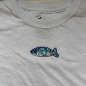 Op de afbeelding: Een wit T-shirt met een kralen visontwerp. De vis is voornamelijk blauw, turquoise en zilver, met een gedetailleerd, getextureerd uiterlijk. Het shirt is effen en heeft een ronde hals.