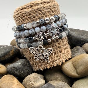 May include: A collection of beaded bracelets with silver charms. The bracelets have grey, black, and clear beads. Charms include a butterfly and a star. The bracelets are displayed on a hessian roll, surrounded by smooth stones.