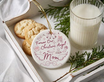 Op de afbeelding: Een witte keramische kerstversiering met de tekst "Forever Our Christmas Angel Mom" in bordeauxrood, versierd met feestelijke illustraties. De versiering hangt boven een dienblad met koekjes en een glas melk, versierd met groen.