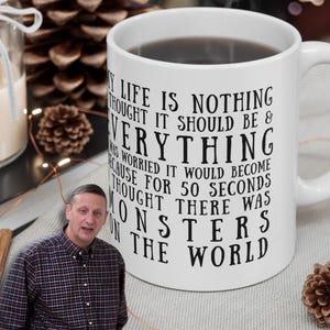 May include: White ceramic mug with black text that reads "My life is nothing thought it should be & everything..." The mug is filled with a dark liquid. The mug is on a table with pinecones and a candle.