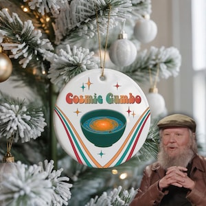 Könnte beinhalten: Ein runder, weißer Keramik-Weihnachtsbaumschmuck mit dem Schriftzug Cosmic Gumbo in bunten Retro-Buchstaben über einer Grafik einer dunkelgrünen Schüssel, die eine wirbelnde orangefarbene Galaxie enthält, akzentuiert durch bunte Linien.