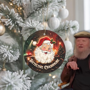 Könnte beinhalten: Ein runder Weihnachtsbaumschmuck aus Keramik mit einer Grafik eines zwinkernden Weihnachtsmanns vor einem dunklen, feurigen Hintergrund mit einem Polizeiauto und dem Schriftzug "DETECTIVE CRASHMORE" in fetter, stilisierter Schrift.