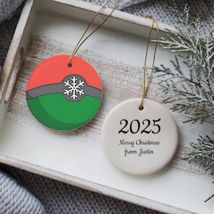 May include: Two round Christmas ornaments. One is red, gray, and green with a snowflake design. The other is white with the year "2025" and "Merry Christmas from Justin" printed on it. Both have gold string hangers.