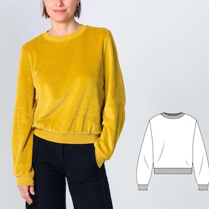 Könnte beinhalten: Ein senfgelbes Langarm-Sweatshirt aus weichem, samtartigem Stoff mit Rundhalsausschnitt sowie gerippten Bündchen an Ärmeln und Taille. Neben dem Kleidungsstück befindet sich eine schwarz-weiße Strichzeichnung, die das Design von vorne zeigt.