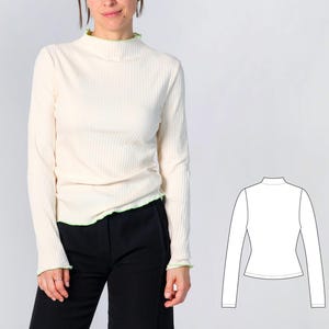 May include: A long-sleeved, ribbed mock neck top in off-white with a light green lettuce-edge trim at the collar, cuffs, and hem. The garment is shown alongside a black and white technical line drawing illustrating the front view of the shirt.