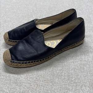 May include: A pair of black leather espadrille flats. The shoes have a rounded toe and a woven jute sole. The interior of the shoe has a gold-colored logo. The shoes are designed for casual wear.