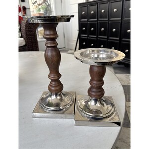May include: Two decorative candle holders. Each has a silver-colored square base, a dark brown wooden stem, and a silver-colored top. The candle holders are on a white surface.