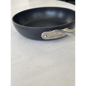 May include: A black non-stick frying pan with a silver handle. The pan has a rounded shape and a slightly textured surface. The handle is attached with rivets and has the brand name "All-Clad" engraved on it. The pan is sitting on a white surface.