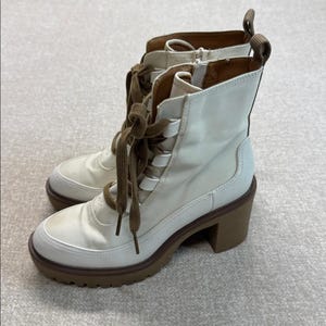 May include: A pair of off-white ankle boots with brown laces and a chunky heel. The boots have a round toe and a lug sole. The laces are threaded through metal eyelets. The heel is approximately 3 inches high.