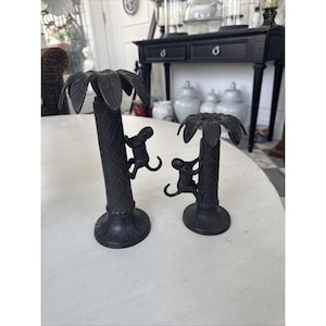 May include: Two black metal candle holders shaped like palm trees, each with a monkey climbing the trunk. The palm fronds form the top of the candle holders. The bases have a decorative, scalloped design.