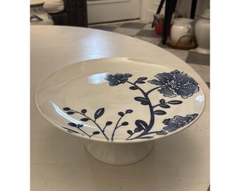 May include: A white ceramic cake stand with a wide, slightly curved top and a short, pedestal base. The top is decorated with a blue floral pattern. The stand is approximately 10 inches in diameter.