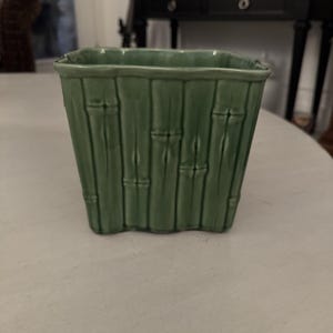 May include: A square, green ceramic planter with a bamboo design. The planter has a textured surface with vertical lines and horizontal bands, mimicking bamboo stalks. The planter is empty and sits on a light-colored surface.