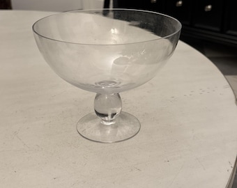 May include: A clear glass dessert bowl with a rounded bowl shape, a small, round stem, and a circular base. The bowl is sitting on a white surface.