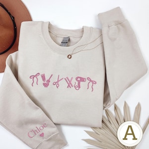 May include: A beige crewneck sweatshirt with pink embroidered salon-themed icons, including scissors and a hairdryer. The left sleeve has the name "Chloe" embroidered in pink with a heart. A gold necklace is draped on the sweatshirt.