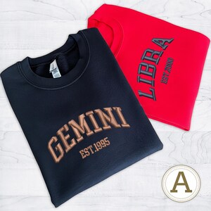May include: Two crewneck sweatshirts, one navy blue and one red. The navy sweatshirt features "GEMINI EST.1995" in a brown, embroidered font. The red sweatshirt displays "LIBRA EST.2000" in a black, embroidered font. Both are on a white wood-grain surface.