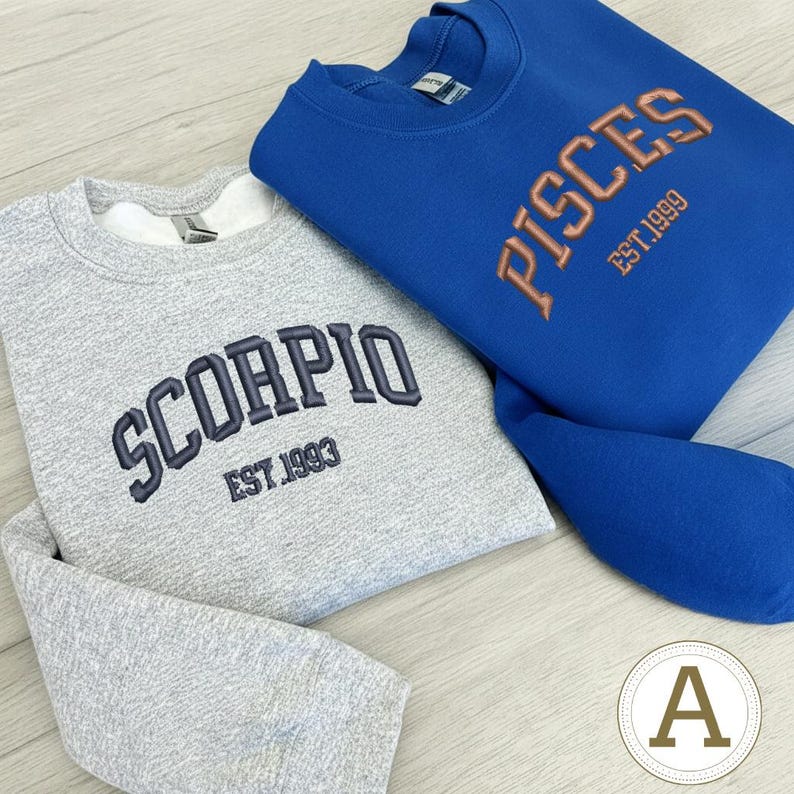 May include: Two sweatshirts are shown, one grey and one blue. The grey sweatshirt has "SCORPIO EST. 1993" embroidered in navy blue. The blue sweatshirt features "PISCES EST. 1999" in orange. Both are crew neck style.