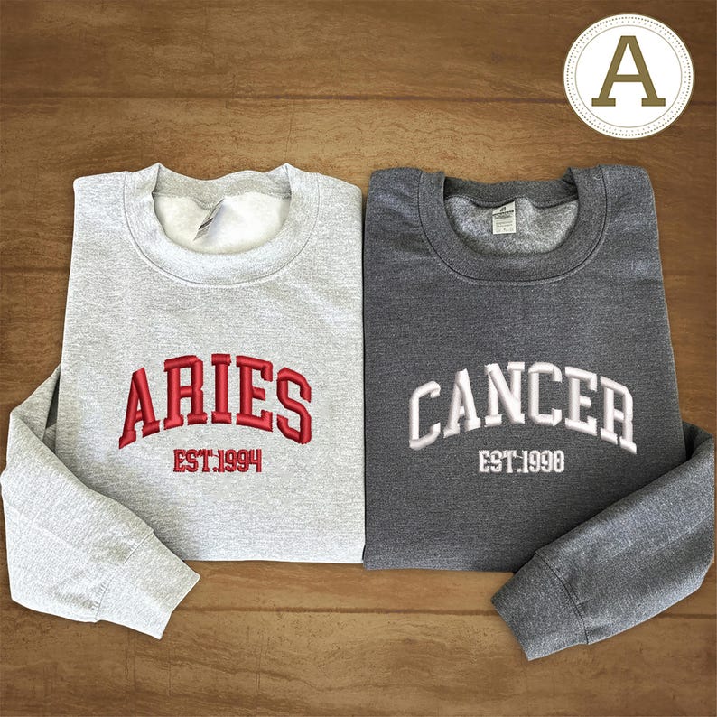 May include: Two crew neck sweatshirts. One is light grey with "ARIES EST.1994" embroidered in red. The other is dark grey with "CANCER EST.1998" embroidered in white. Both are folded on a wooden surface.