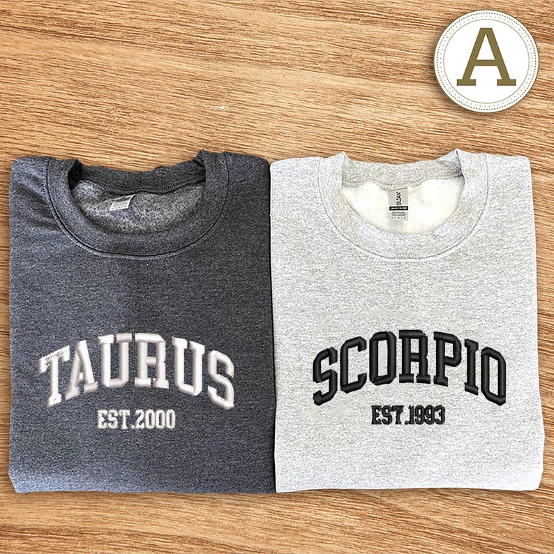 May include: Two crew neck sweatshirts, one dark grey and one light grey. The dark grey sweatshirt has "TAURUS" and "EST. 2000" embroidered in white. The light grey sweatshirt has "SCORPIO" and "EST. 1993" embroidered in black.
