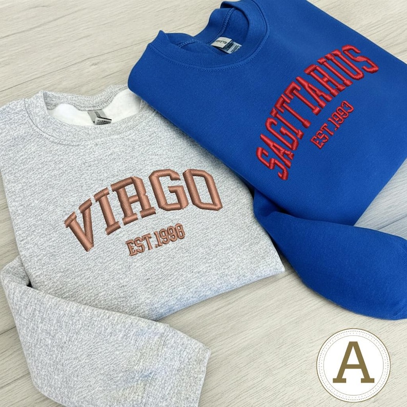 May include: Two crew neck sweatshirts, one grey and one blue. The grey sweatshirt has "VIRGO EST 1998" embroidered in copper. The blue sweatshirt has "SAGITTARIUS EST 1993" embroidered in red. Both have a simple, classic design.