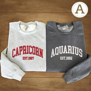 May include: Two grey sweatshirts with embroidered text. One is light grey with "CAPRICORN EST.1997" in red. The other is dark grey with "AQUARIUS EST.1992" in white. Both have a crew neck and long sleeves.